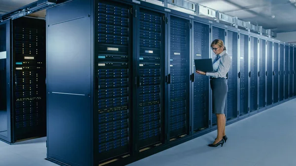 Em Data Center: Técnico de TI fêmea fica perto do gabinete da cremalheira e executa o programa de manutenção no laptop, controla o ótimo funcionamento do servidor operacional . — Fotografia de Stock