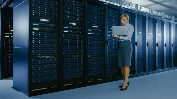 In Data Center: Female IT Technician Stands Near Rack Cabinet and Runs Maintenance Program on Laptop, Controls Operational Server\'s Optimal Functioning.