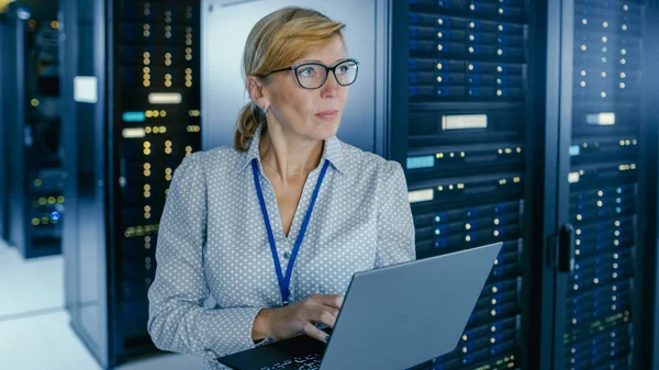 Portrait Shot In Data Center: Female IT Technician Running Maintenance Programme on a Laptop, Controls Operational Server Rack Optimal Functioning. High-Tech Telecommunications Operational Data Center