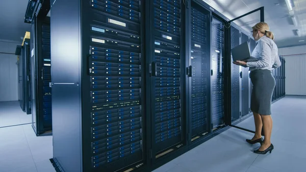 En el centro de datos: Técnico de TI femenino se para cerca del gabinete de rack abierto y ejecuta el programa de mantenimiento en el ordenador portátil, controla el funcionamiento óptimo del servidor operativo . — Foto de Stock