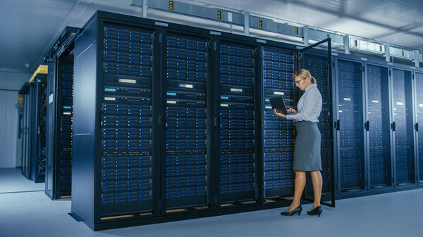 In Data Center: Female IT Technician Stand Near Opened Rack Cabinet and Runs Maintenance Program on Laptop, Controls Operational Server's Optimal Functioning.