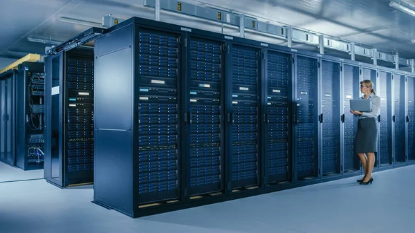 Em Data Center: Especialista em TI Feminina Caminha ao longo da Linha de Racks de Servidores Operacionais, Utiliza Laptop para Executar Programa de Manutenção. Moderno Data Center Operacional de Telecomunicações de Alta Tecnologia . — Fotografia de Stock