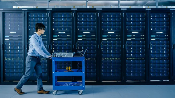 No Data Center Moderno: Engenheiro de TI Empurra Carrinho ao lado de Racks e Gabinetes de Servidor para Instalar Novo Hardware para Atualização Planejada do Sistema, Substituição de Equipamentos . — Fotografia de Stock