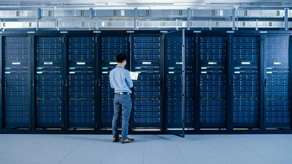 No centro de dados moderno: O coordenador de TI que está ao lado dos armários abertos da cremalheira do servidor, faz a manutenção sem fio e o procedimento dos diagnósticos com um portátil . — Fotografia de Stock