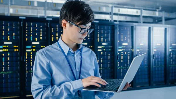 En el centro de datos moderno: Retrato del ingeniero de TI parado con bastidores del servidor detrás de él, haciendo el mantenimiento y el procedimiento de diagnóstico usando el ordenador portátil . — Foto de Stock