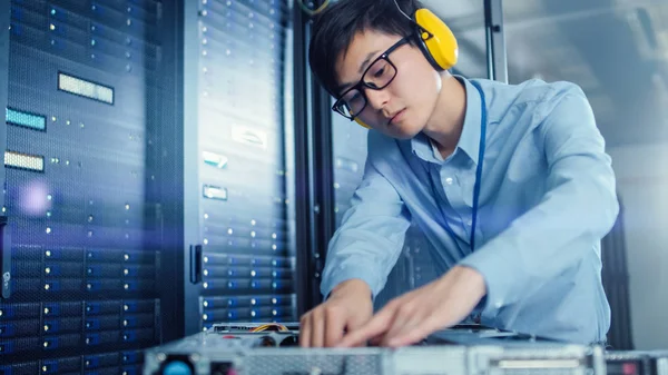 In the Modern Data Center: IT Engineer Wearing Protective Headphones Installs New Hardware for Server Rack. IT Specialist Doing Maintenance, Running Diagnostics and Updating Hardware.