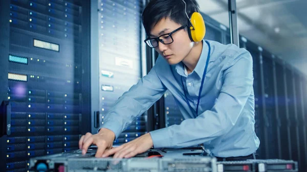 In the Modern Data Center: IT Engineer Wearing Protective Headphones Installs New Hardware for Server Rack. IT Specialist Doing Maintenance, Running Diagnostics and Updating Hardware.