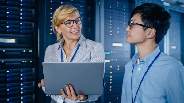 In the Modern Data Center: Engineer and IT Specialist Walking alongside Server Racks, Talking, Doing System Maintenance Check with Laptop Computer. Specialists Doing Diagnostics of the Database.