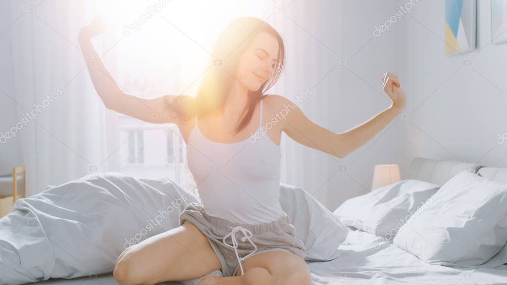 Beautiful Brunette is Waking up in the Morning, Stretches in the Bed, Sun Shines on Her From the Big Window. Happy Young Girl Greets New Day with Warm Sunlight Flare.