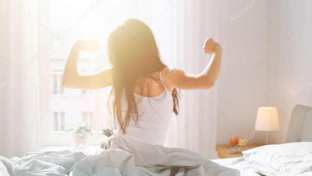 Beautiful Brunette is Waking up in the Morning, Stretches in the Bed, Sun Shines on Her From the Big Window. Happy Young Girl Greets New Day with Warm Sunlight Flare.