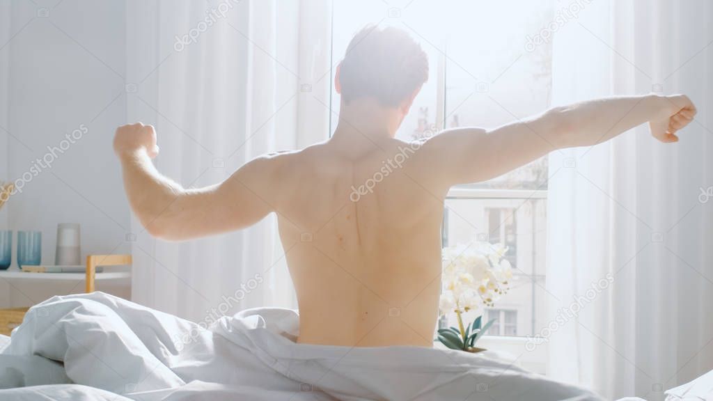 Handsome Fair Boy Wakes up after Sleeping, Gets out of Bed and Stretches Lazily in a Bed. Young Caucasian Man. Early Morning Sun Shines Through the Window