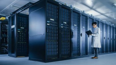 Veri Merkezi: erkek o uzman beyaz ceket giyen sunucu raf yanında standları, bakım tanılama araçları çalıştırmak için dizüstü bilgisayar kullanır, böylece veritabanı En Iyi düzeyde çalışır kontrol.