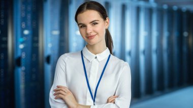Female It Specialist, sunucu rafları ve kameraya bakarak veri merkezinde kamera 'da duruyor. Onun Arms çapraz ve o kamera Smiles.