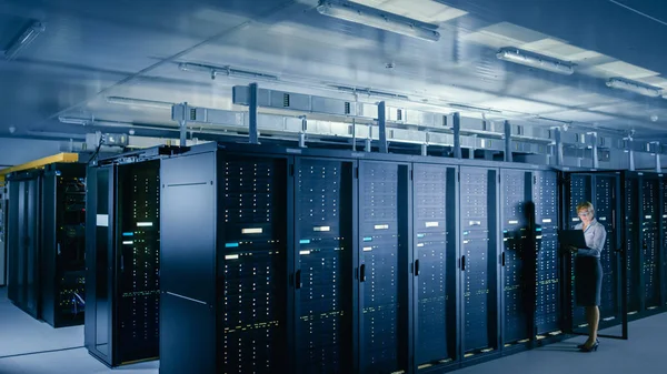 W centrum danych: Kobieta IT technika stoi przed Open Server rack gabinetu, używa laptopa do uruchomienia diagnostyki konserwacji tak, że mainframe pracuje w: optimum funkcjonowanie Level. — Zdjęcie stockowe