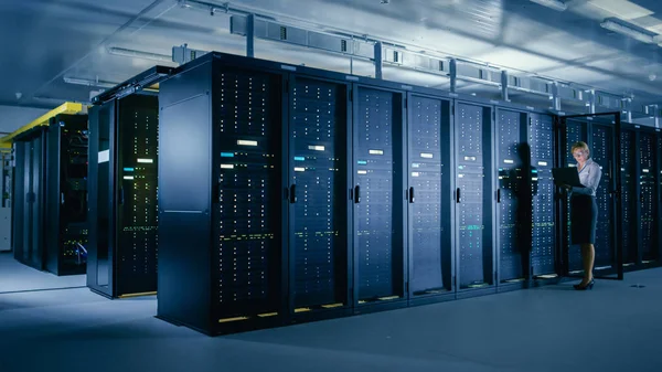 W centrum danych: Kobieta IT technika stoi przed Open Server rack gabinetu, używa laptopa do uruchomienia diagnostyki konserwacji tak, że mainframe pracuje w: optimum funkcjonowanie Level. — Zdjęcie stockowe