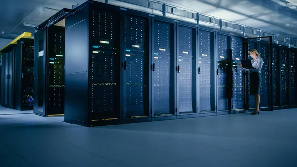データセンター:女性IT技術者がオープンサーバラックキャビネットの前に立ち、ラップトップコンピュータを使用してメンテナンス診断を実行し、メインフレームが最適な機能レベルで動作するように. — ストック写真