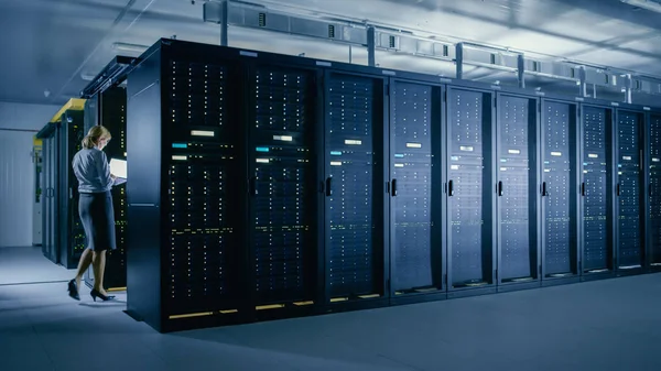 В центре обработки данных: женщина-IT-техник проходит между рядами серверов, использует портативный компьютер для запуска диагностики технического обслуживания, чтобы мэйнфрейм работал на оптимальном уровне функционирования . — стоковое фото