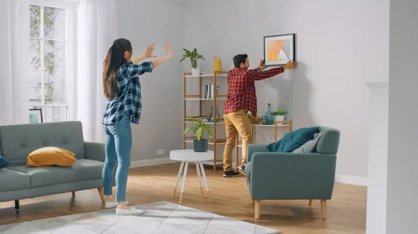 Feliz casal pendurado imagem na parede, namorado move-lo, namorada diz-lhe quando o quadro é pendurado em linha reta. Momento engraçado na vida de um jovem casal. Apartamento moderno e elegante . — Fotografia de Stock