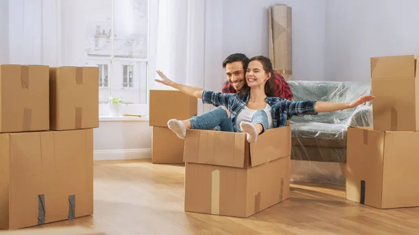 Happy Young Couple Moving Into New Apartment, Boyfriend Drives Girlfriend in Cardboard Box, She imitates Airplane Wings. Young Boyfriend, Girlfriend Start Living Together, Unpacking Stuff, Having Fun.