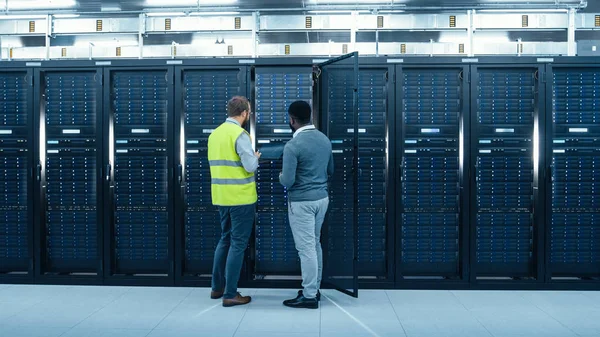 IT administrator med en bærbar computer i høj sigtbarhed Vest og Black Male Engineer taler i datacenter, mens stående foran en server Rack. Kører diagnostik eller udfører vedligeholdelsesarbejde . - Stock-foto