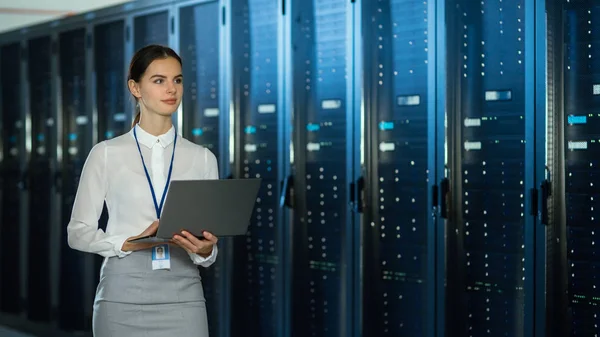 Hermoso centro de datos Técnico de TI femenino que camina a través del corredor del estante del servidor con una computadora portátil. Ella está inspeccionando visualmente los gabinetes del servidor de trabajo . — Foto de Stock