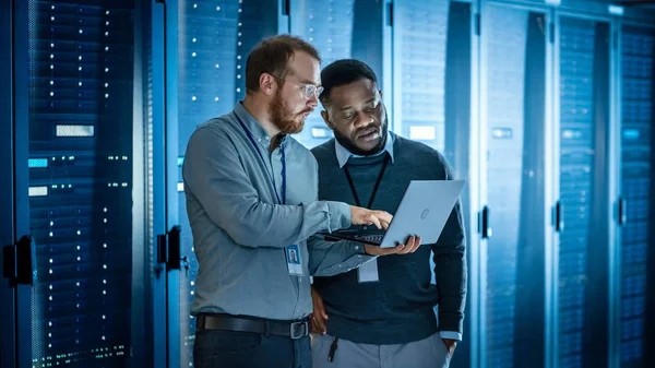 Bearded IT Technician in Glasses with Laptop Computer and Black Male Engineer Colleague are Using Laptop in Data Center while Working Next to Server Racks. Running Diagnostics or Doing Maintenance Work — Stock Photo, Image