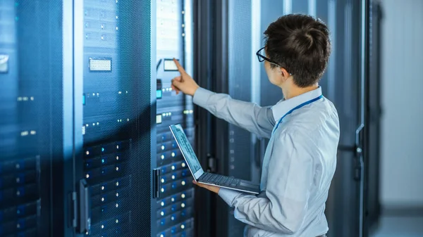 En el centro de datos moderno: Ingeniero de TI junto a gabinetes de bastidor de servidor abierto, hace el mantenimiento inalámbrico y el procedimiento de diagnóstico con un ordenador portátil. Él presiona el botón . — Foto de Stock
