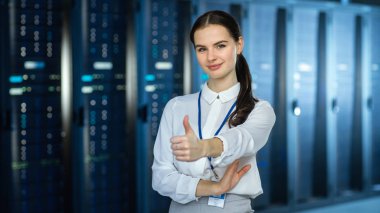 Female It Specialist, sunucu rafları ve kameraya bakarak veri merkezinde kamera 'da duruyor. O Thumb yukarı ve kamera Smiles gösterir.