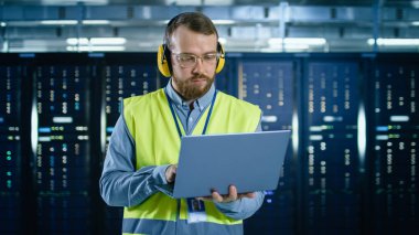 Gözlük ve kulaklıklarda sakallı IT uzmanı, yüksek görünürlük yelek giyen sunucu rafları yanında veri merkezi dizüstü bilgisayarda çalışıyor.