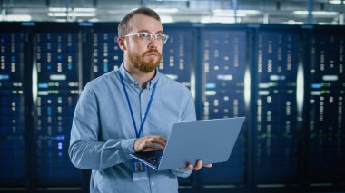 Gözlüklerin sakallı IT uzmanı, sunucu rafları 'nın yanındaki veri merkezinde dizüstü bilgisayarda çalışıyor. Tanılama çalıştırma veya bakım çalışması yapma.