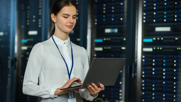 Hermoso centro de datos Técnico de TI femenino de pie cerca del corredor del estante del servidor con una computadora portátil. Ella está usando Notebook y trabajando con servidores . — Foto de Stock