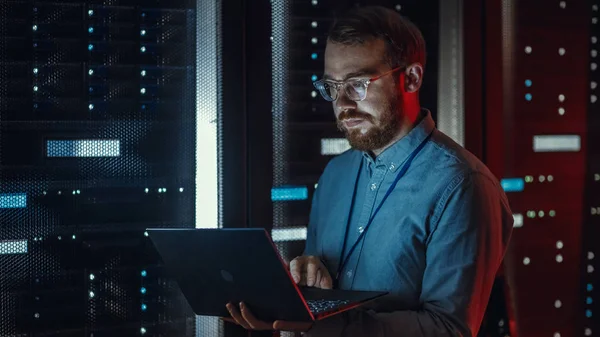 Бородатый IT-специалист по очкам работает над ноутбуком в дата-центре, стоя рядом с серверной стойкой. Провожу диагностику, техническое обслуживание. Аварийный красный свет от специалиста по светильникам сбоку — стоковое фото