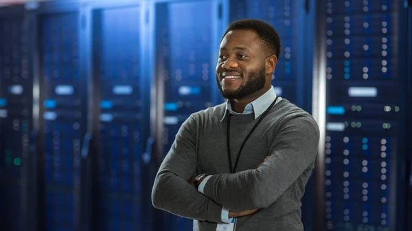 Black Data Center IT Technician Standing in the Middle of a Server Rack Corridor. He is Standing with Crossed Arms and is Smiling.