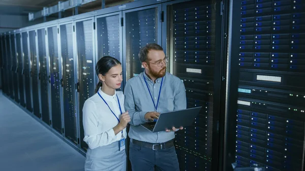 Skäggiga IT-tekniker i glas med en bärbar dator och vackra unga ingenjör kollega pratar i Data Center medan du arbetar bredvid Server rack. Köra diagnostik eller utföra underhållsarbete. — Stockfoto