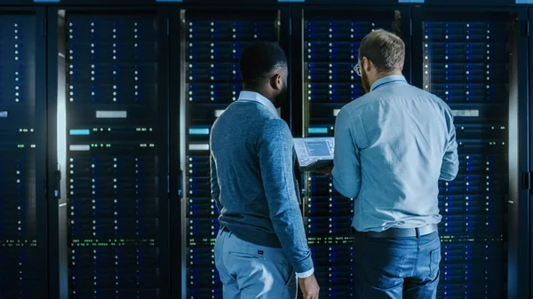 Bearded IT-technicus in glazen met een laptop computer en zwarte mannelijke ingenieur collega werken in Data Center tijdens het werken naast server racks. Het uitvoeren van diagnostische gegevens of onderhoudswerkzaamheden. — Stockfoto