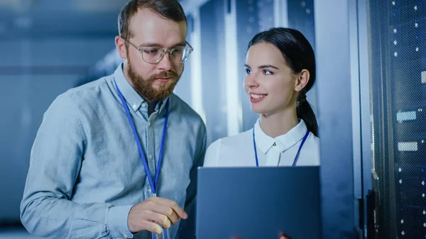 Szakállas it technikus a szemüveg és gyönyörű fiatal mérnök kolléga egy laptop számítógép beszél Data Center mellett munka mellett Server racks. A diagnosztika futtatása vagy karbantartási munka során. — Stock Fotó