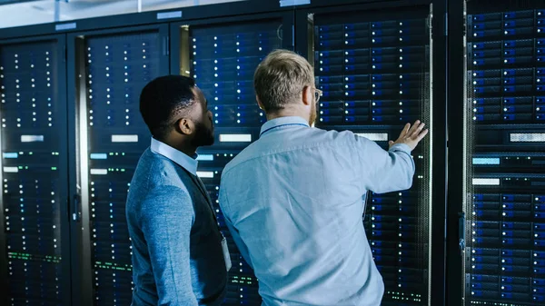 Bir laptop bilgisayar ve siyah erkek mühendisi meslektaşım ile gözlük sakallı IT teknisyeni sunucu rafları yanında çalışırken veri merkezi 'nde konuşuyor. Tanılama çalıştırma veya bakım çalışması yapma.