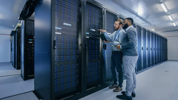 Técnico de TI Barbudo em Óculos com Computador Laptop e Colega de Engenheiro Masculino Preto estão falando no Data Center enquanto trabalham ao lado de Racks de Servidor. Executando diagnósticos ou fazendo trabalhos de manutenção . — Fotografia de Stock