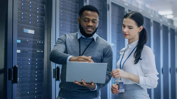 Black IT Technician with a Laptop Computer Gives a Tour to a Young Intern. Hablan en el centro de datos mientras caminan junto a los bastidores del servidor. Ejecución de diagnósticos o trabajos de mantenimiento . — Foto de Stock