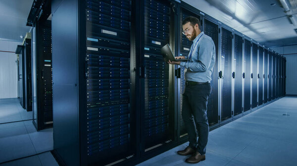 Bearded IT Specialist in Glasses is Working on Laptop in Data Center while Standing Before Server Rack. Running Diagnostics or Doing Maintenance Work.