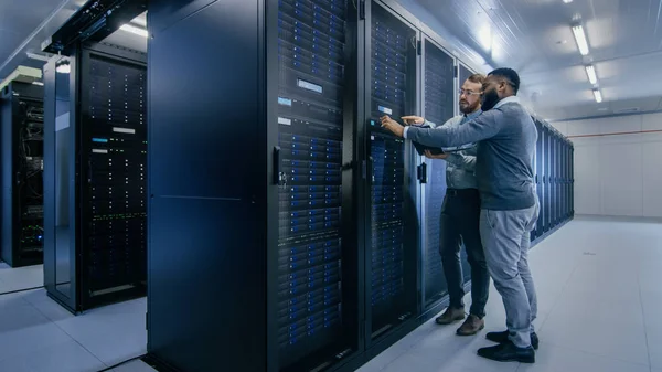 Brodaty IT technika w okularach z laptopem i czarny mężczyzna inżynier kolega rozmawiają w centrum danych podczas pracy obok szafy serwerowe. Uruchamianie diagnostyki lub wykonywanie prac konserwacyjnych. — Zdjęcie stockowe