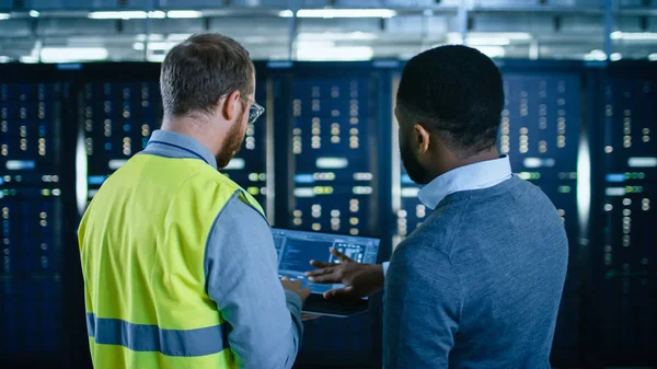 Voltar Visão no Engenheiro de TI Barbudo em Óculos e Colete de Alta Visibilidade com um Computador Laptop e Colega Técnica Preta Falando no Data Center enquanto está ao lado de Racks de Servidor. Executando diagnósticos ou manutenção . — Fotografia de Stock