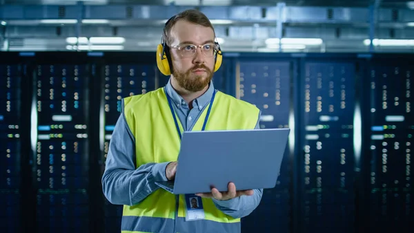 Especialista em TI Barbuda em óculos e fones de ouvido, vestindo colete de alta visibilidade está trabalhando no laptop em data center ao lado de cremalheiras de servidor . — Fotografia de Stock