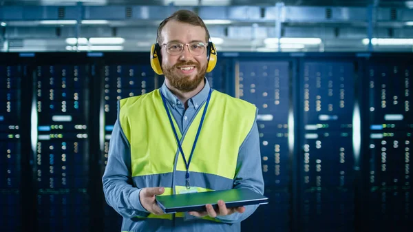 Bärtig mit Brille und Kopfhörer lächelt er mit Warnweste in die Kamera im Rechenzentrum neben Serverschränken. Er hält den Laptop in der Hand. — Stockfoto