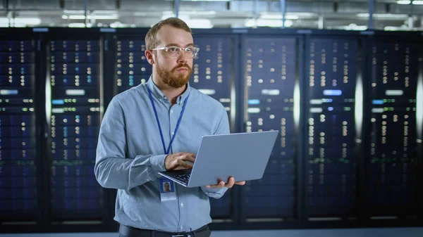 Bearded IT specialist in glazen werkt op laptop in Data Center naast server racks. Het uitvoeren van diagnostische gegevens of onderhoudswerkzaamheden. — Stockfoto