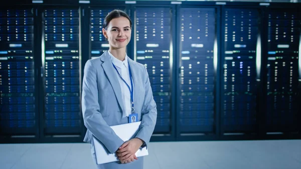 Vrouwelijke IT-specialist staat in Data Center naast server racks, houdt laptop en glimlacht naar de camera. — Stockfoto