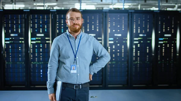 Gözlüklere sakallı IT uzmanı, sunucu rafları yanında, kamera ve holdıng laptop için gülümseyen veri merkezi 'nde ayakta. Tanılama çalıştırma veya bakım çalışması yapma.