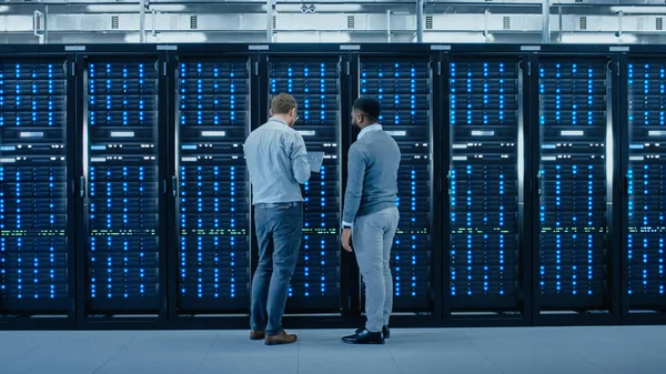 Bearded IT Engineer in Glasses with a Laptop Computer and Black Technician Colleague Talking in Data Center while Standing Next to Server Racks. Running Diagnostics or Maintenance. — Stock Photo, Image