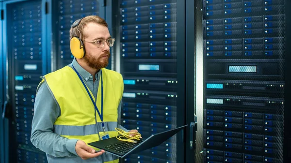 Bearded IT Specialist in Glasses and Headphones, wearing High Visibility Vest is Working on Laptop in Data Center Next to Server Racks. Running Diagnostics or Doing Maintenance Work. — Stock Photo, Image