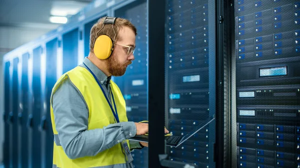 IT Engineer in High Visibility Vest is Working on Laptop in Data Center while Standing Before Server Rack. Running Diagnostics or Doing Maintenance Work.